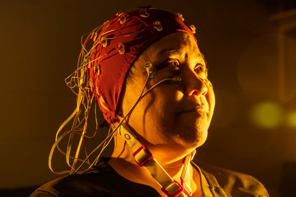 A person wearing a red EEG cap with attached electrodes sits in warm, dramatic lighting. Their calm expression suggests focus and concentration.