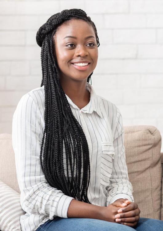A woman with long braided hair smiles warmly, seated on a beige sofa against a white brick wall. She wears a striped shirt, exuding a relaxed and cheerful vibe.